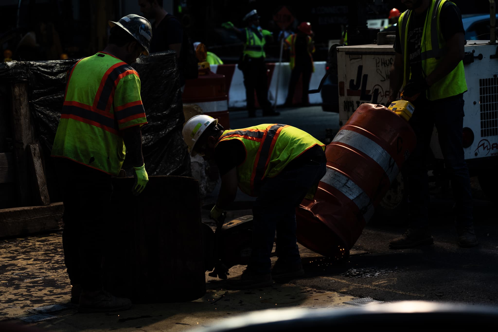 Construction workers in high-visibility vests are engaged in work near a vehicle, with one bending over what appears to be a barrel or drum. The scene is dimly lit, suggesting either an evening setting or a shaded area