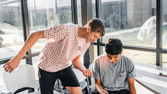 Two individuals in a modern office setting, one standing and leaning over to look at the other's computer screen, possibly collaborating or discussing a project