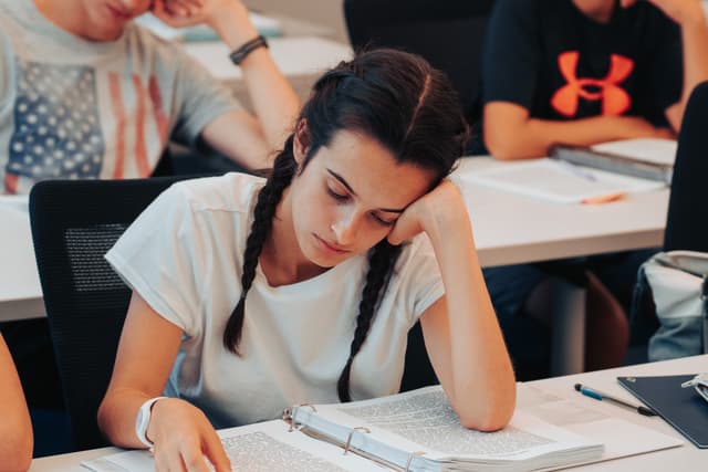 A focused young woman with braided hair is studying in a classroom, leaning on her hand with notebooks and pens on the desk in front of her. Other students are visible in the background