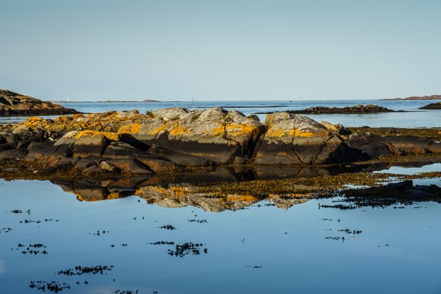 A serene coastal landscape with rocks reflected in calm water under a clear blue sky