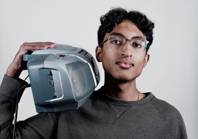 A young individual with glasses, wearing a dark long-sleeved shirt, holds a virtual reality headset beside their head, looking at the camera with a slight smile