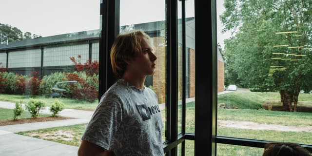 Emiliano Garcia-Lopez, looking outside with a contemplative expression, in a room that appears to be a lobby or common area with a view of greenery and a building outside