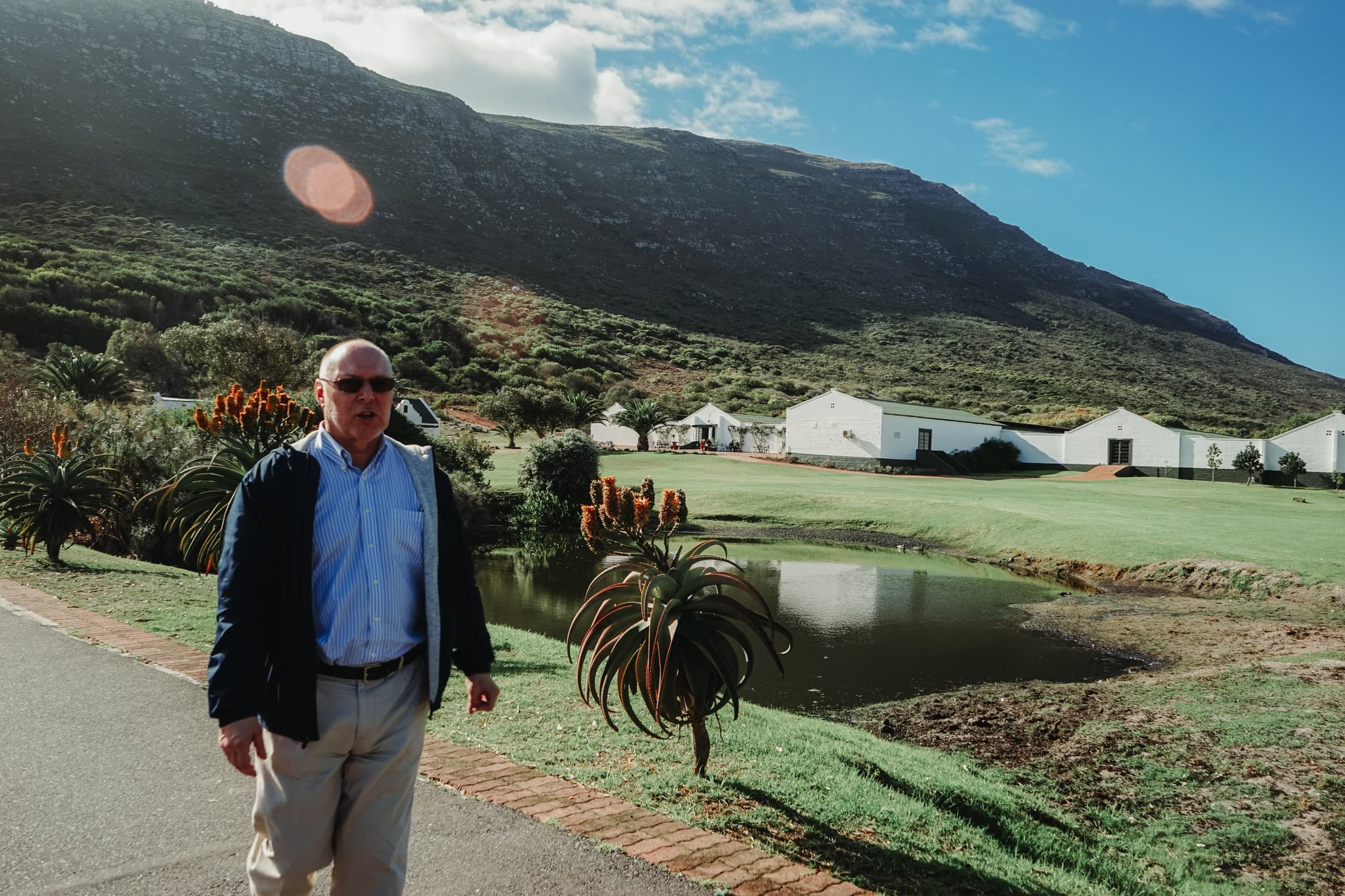 A man stands on a road with a mountainous backdrop, a clear sky, and a body of water reflecting the surrounding greenery