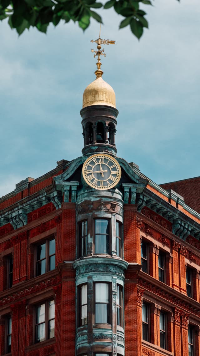 A red brick building with a distinctive clock tower topped by a golden dome and weathervane, framed by a blue sky and partially obscured by green foliage