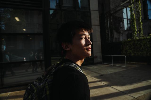 A young man with a backpack stands in a sunlit area, with shadows casting across his face and the building behind him