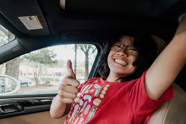 A person in a red shirt is giving a thumbs-up while sitting in the driver's seat of a car, smiling broadly