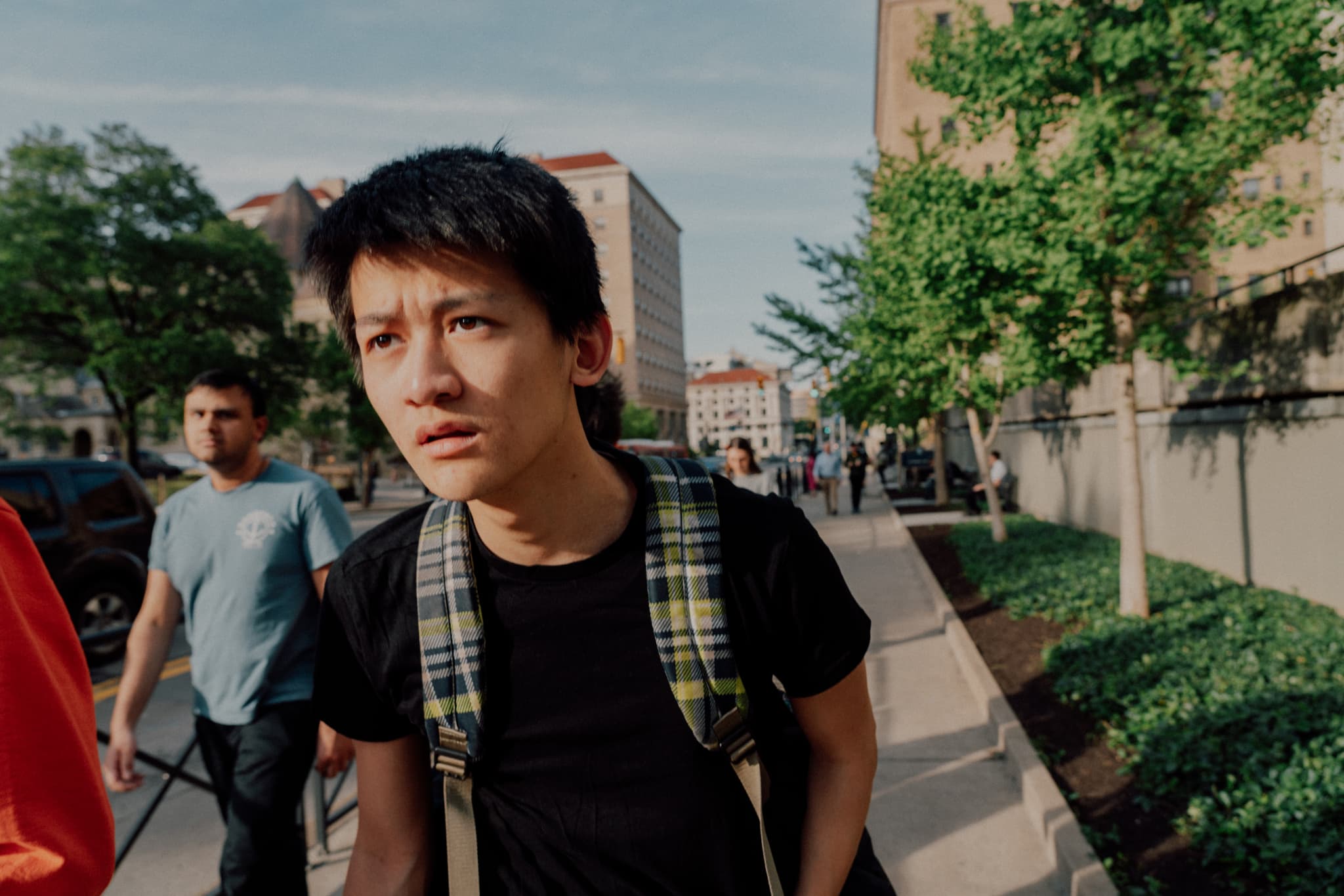 A young person with a backpack looks concerned while walking on a city sidewalk, with other pedestrians and buildings in the background