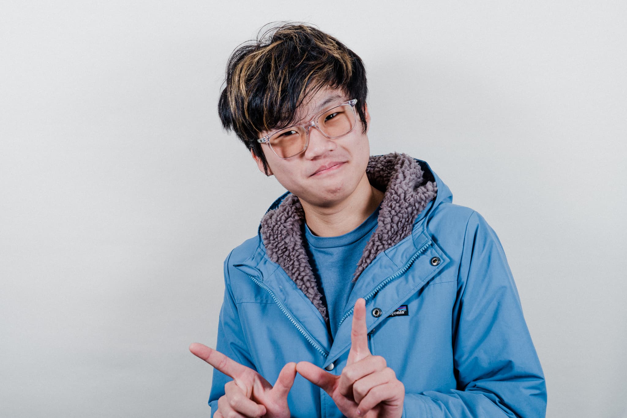 A person with tousled hair wearing glasses and a blue jacket over a patterned shirt, making a playful expression and gesture with their fingers crossed in front of them