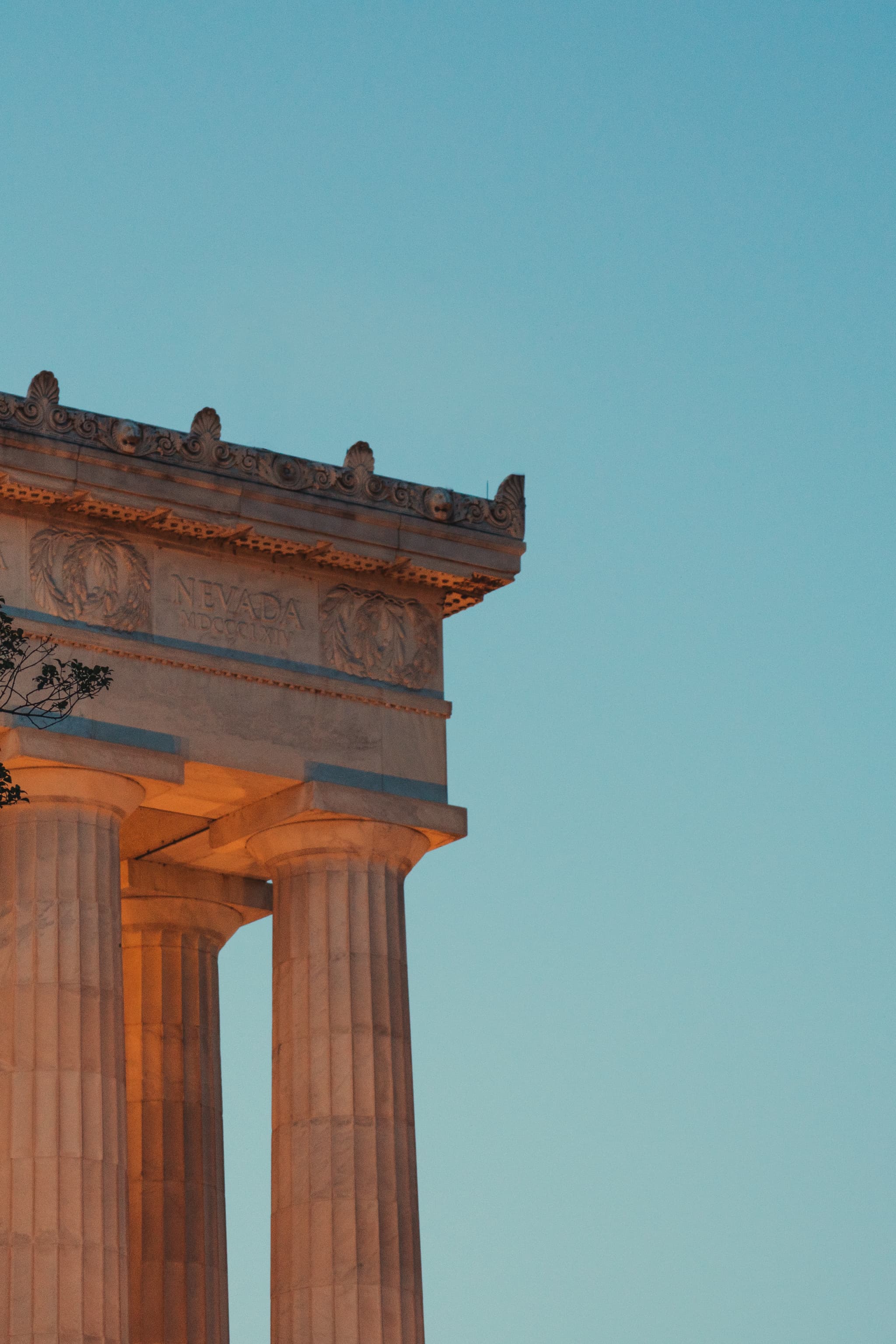 A section of a classical columned structure against a clear blue sky during what appears to be dusk or dawn, with sunlight illuminating the columns