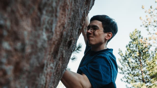 A man wearing glasses is rock climbing, gripping the rock face with focus and determination