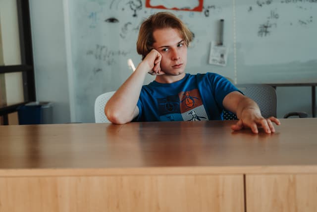 Emiliano Garcia-Lopez, with a contemplative expression is resting his head on his hand at a wooden table, with a whiteboard in the background