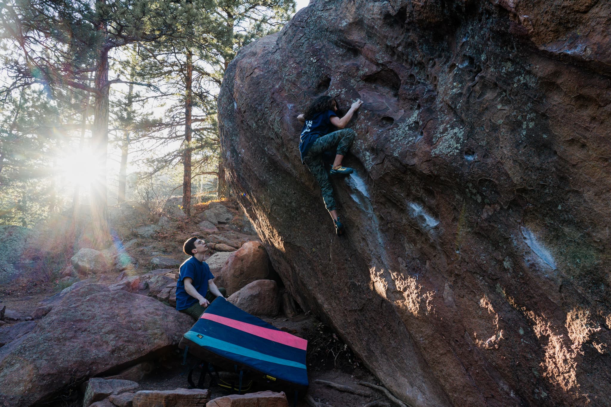 A climber grips a large boulder, attempting to navigate its surface, while another person stands below with a crash pad, ready to assist. Sunlight filters through the trees, adding a warm glow to the scene