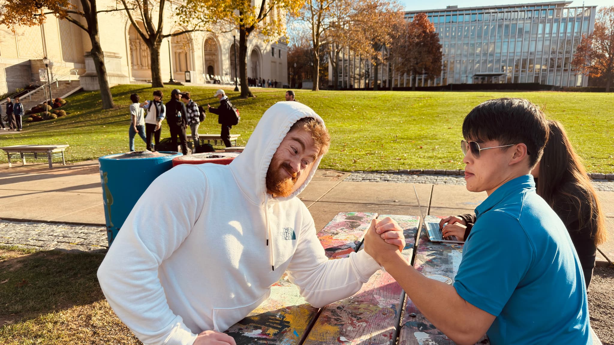 Two individuals are seated at an outdoor table, one making a playful face at the camera while the other looks on. They appear to be engaged in a casual, social setting with a clear sky and buildings in the background