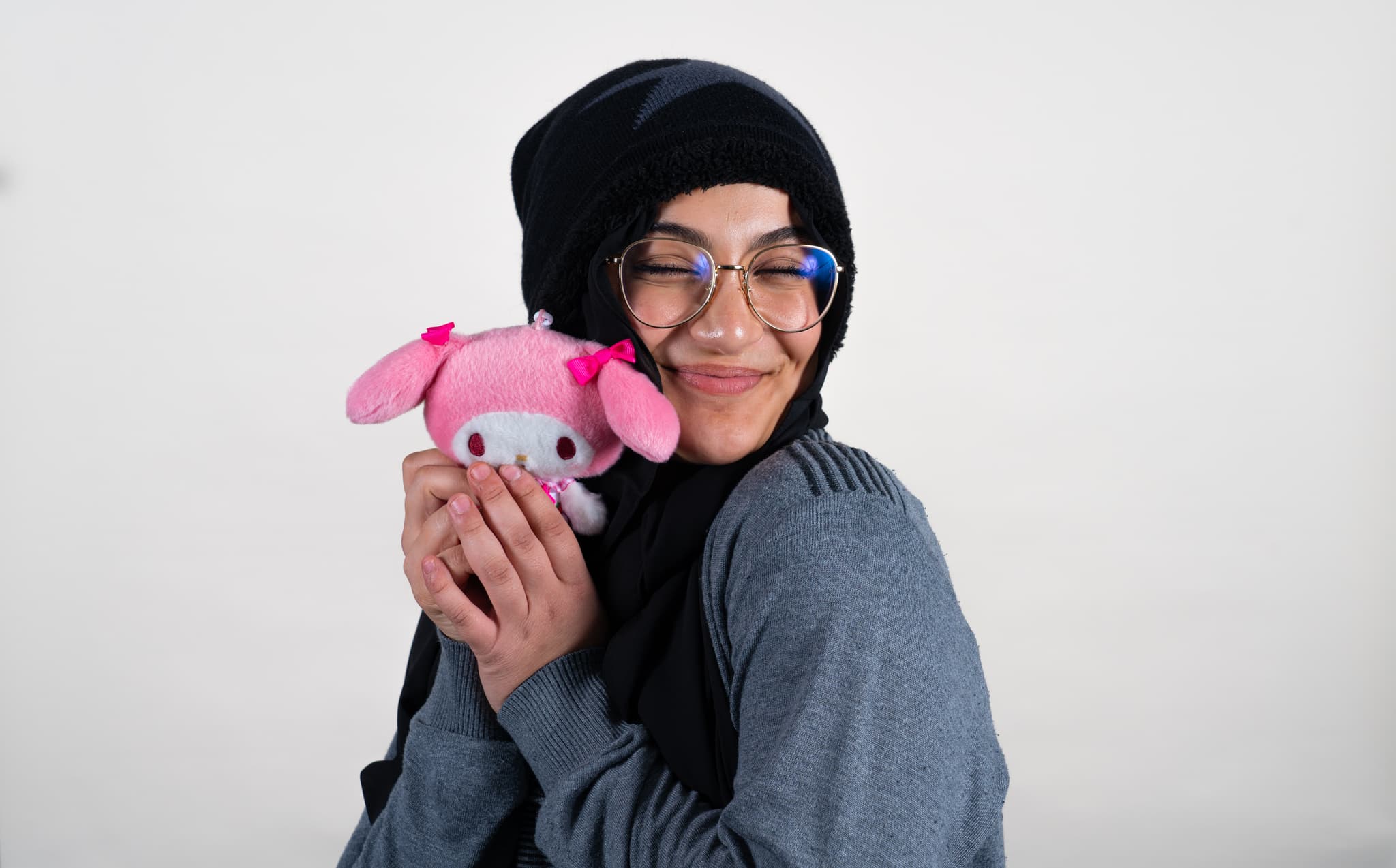 A smiling person wearing glasses and a black headscarf hugs a plush pink pig toy against a plain background