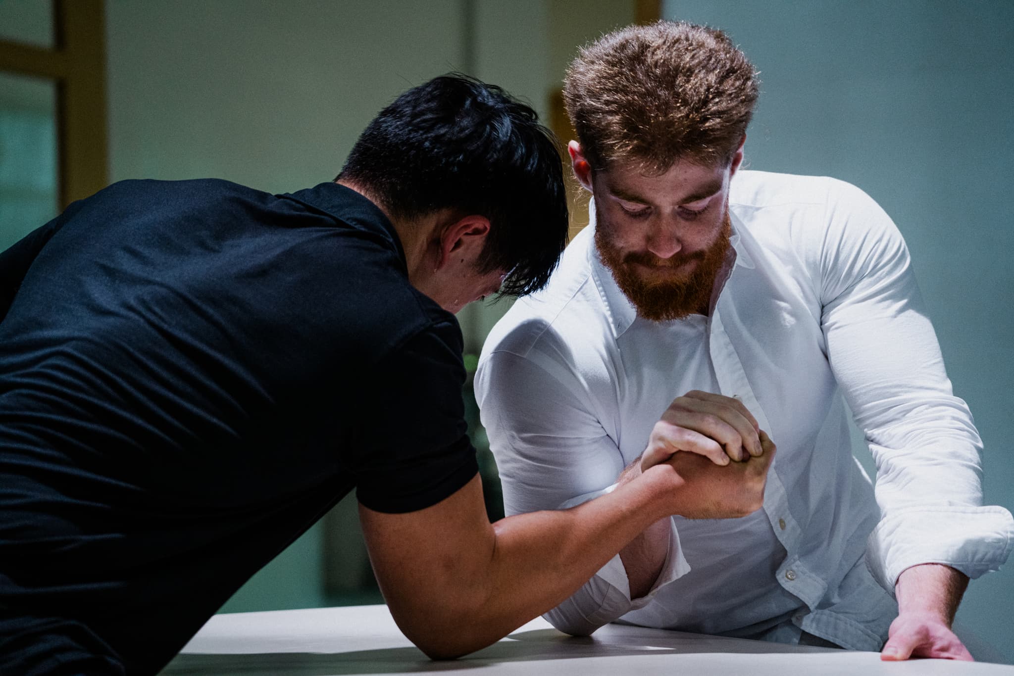 Two men engaged in an arm wrestling match, one wearing a white shirt and the other in black, both concentrating intensely on the contest