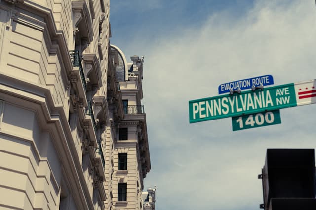 A street sign reading Pennsylvania Ave NW 1400 against a backdrop of a building and a partly cloudy sky