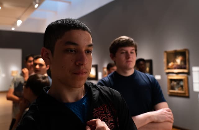 Two young men stand in the foreground at an art gallery, with one looking towards the camera and the other looking away, while other visitors and artwork are visible in the background