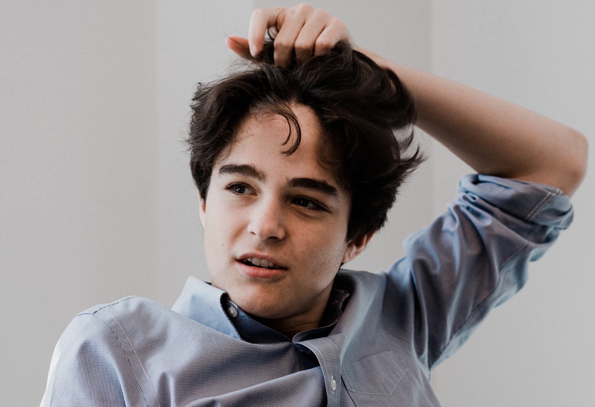 A young individual with dark hair wearing a blue shirt is running their fingers through their hair, looking slightly to the side with a thoughtful expression