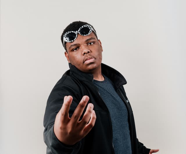 A person wearing a headband and a black jacket extends their hand towards the camera, with a neutral expression on their face, against a plain background