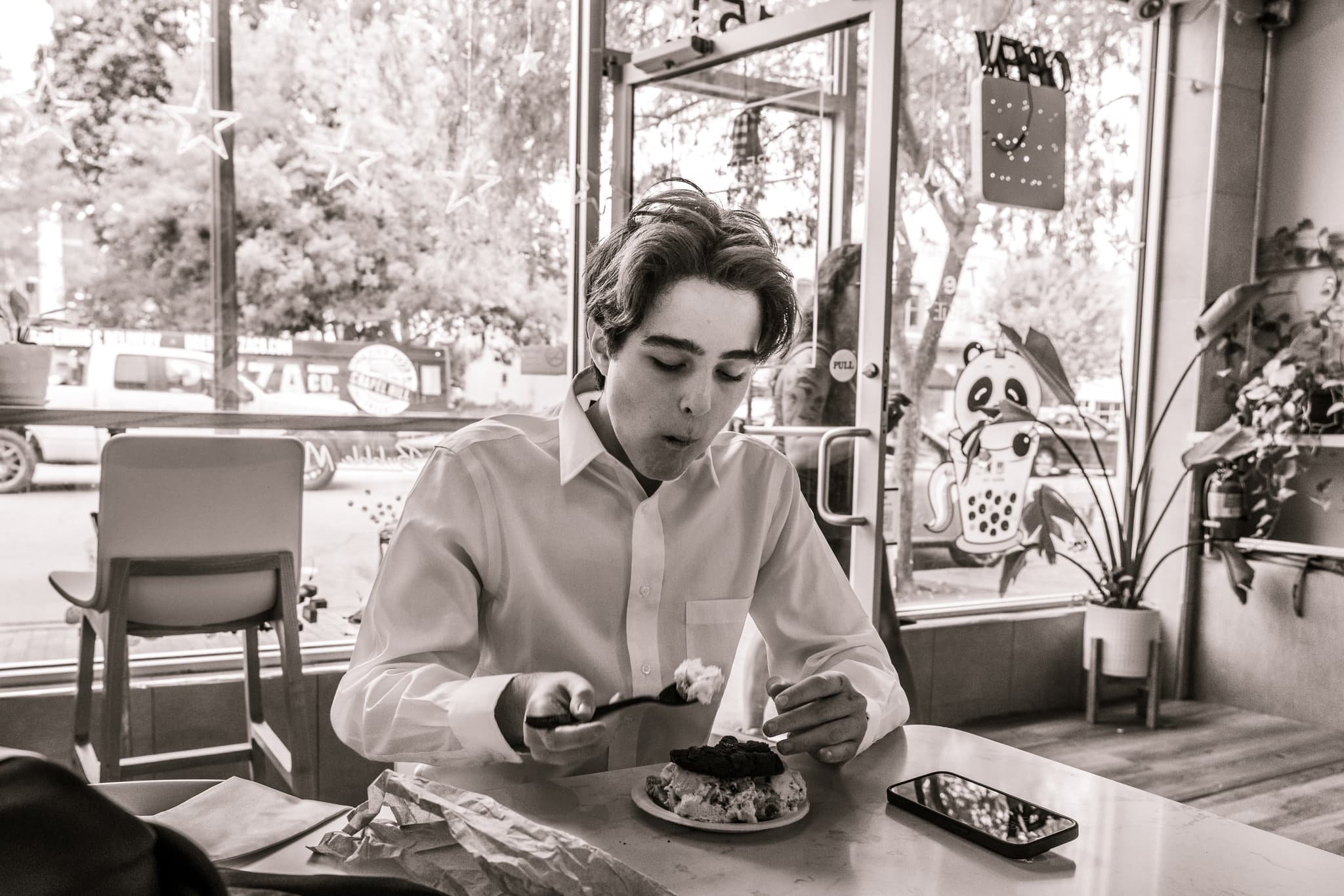 A black and white photo of a person sitting at a cafe table, looking at their phone with a plate of food in front of them