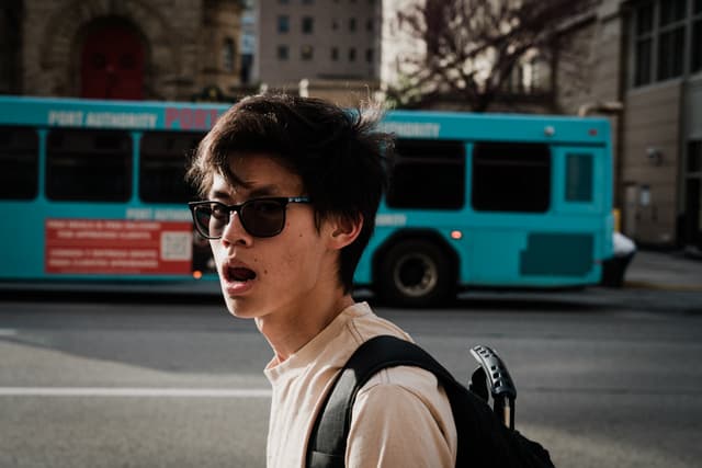 A surprised young man wearing sunglasses and a backpack on a city street with a blue bus in the background