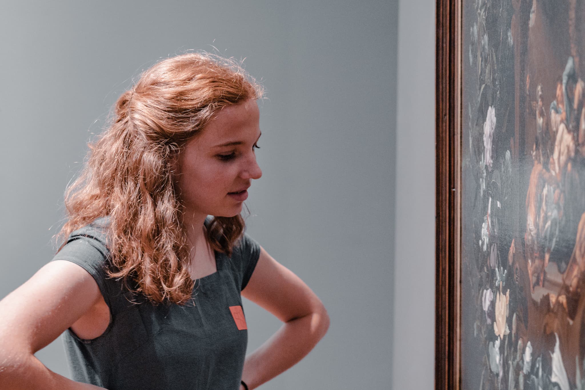 A woman with curly hair, wearing a sleeveless top, is looking intently at a painting in an art gallery