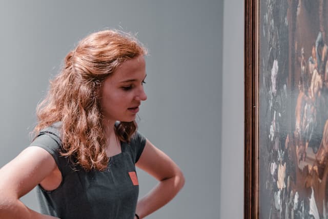 A woman with curly hair, wearing a sleeveless top, is looking intently at a painting in an art gallery