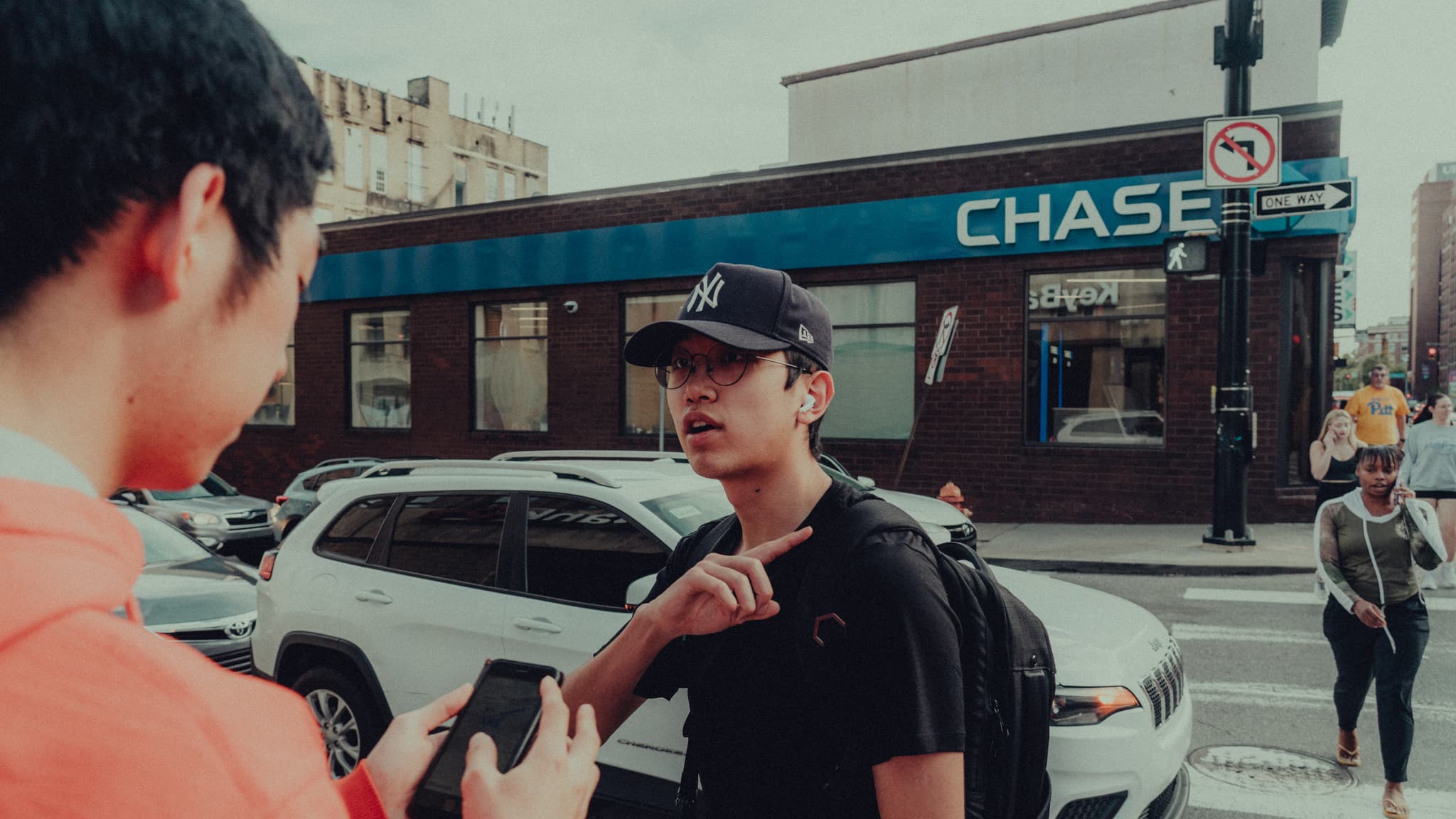 Two individuals are engaged in a conversation on a city street, with one person holding a smartphone and gesturing towards the other, who is listening attentively. In the background, there's a Chase bank branch and urban traffic