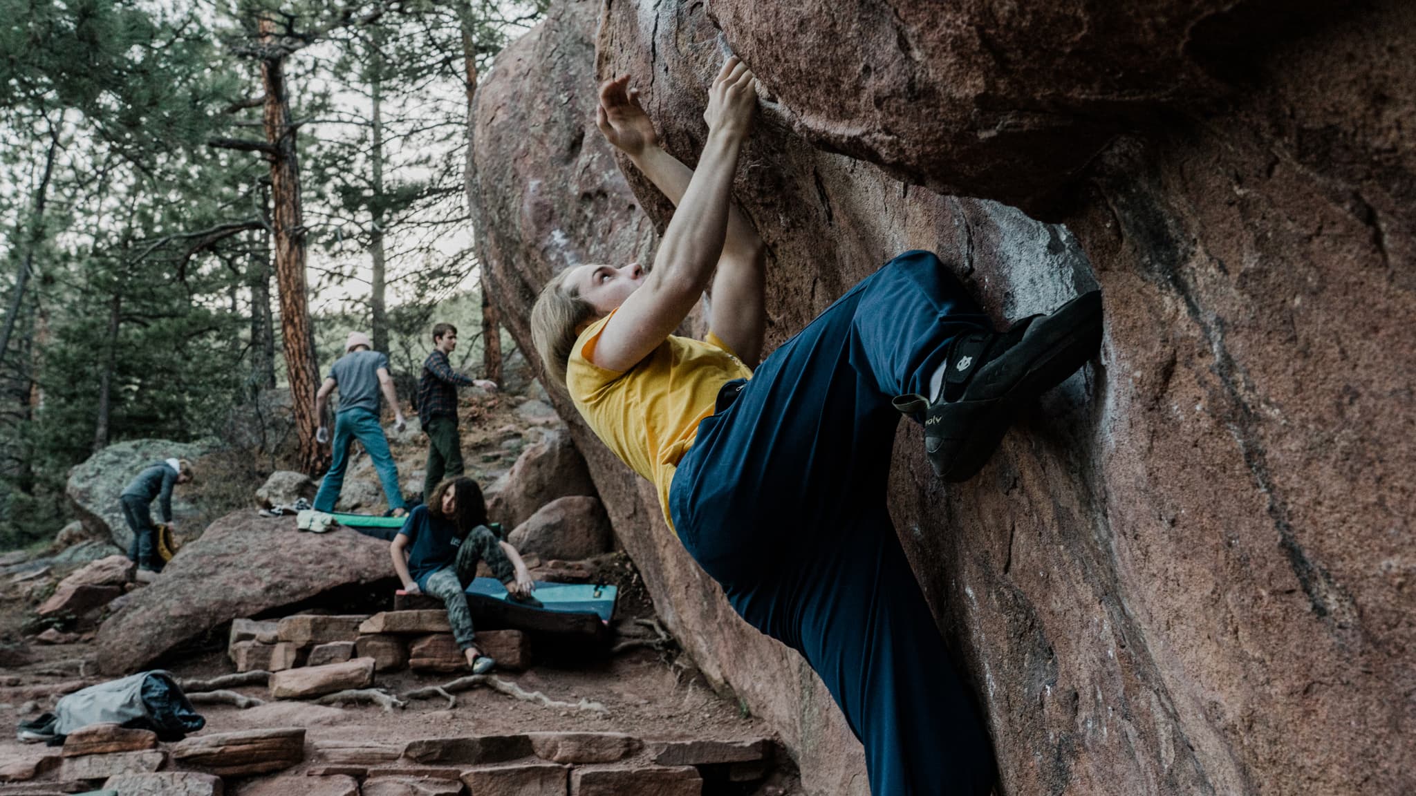 A climber grips a boulder with crash pads below, while another person watches from the background amidst a wooded area