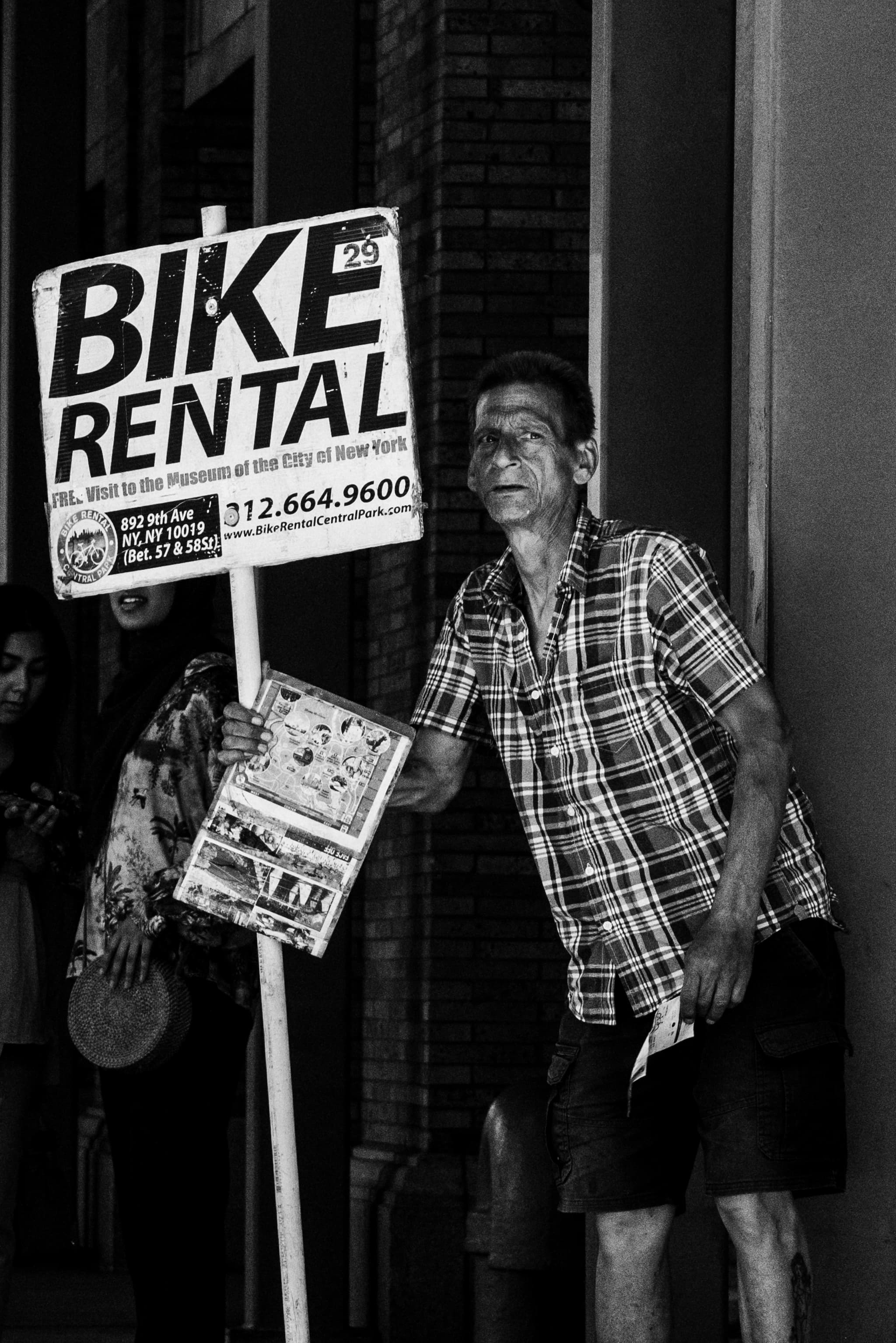 A black and white photo capturing a man standing on a sidewalk holding a BIKE RENTAL sign, with a newspaper tucked under his arm