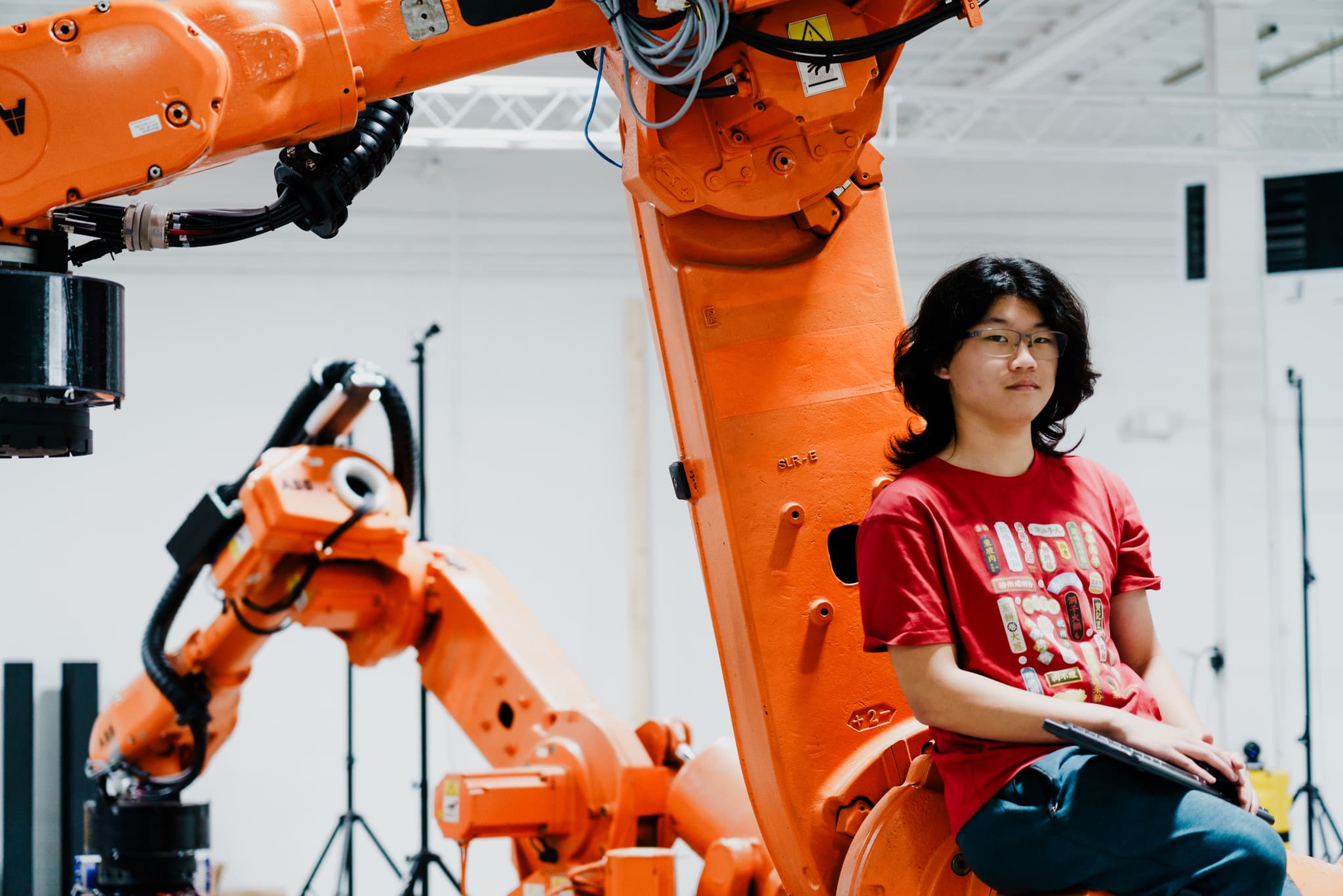 A person sits in front of an industrial robot, appearing contemplative or focused, in a setting that suggests a workshop or robotics lab