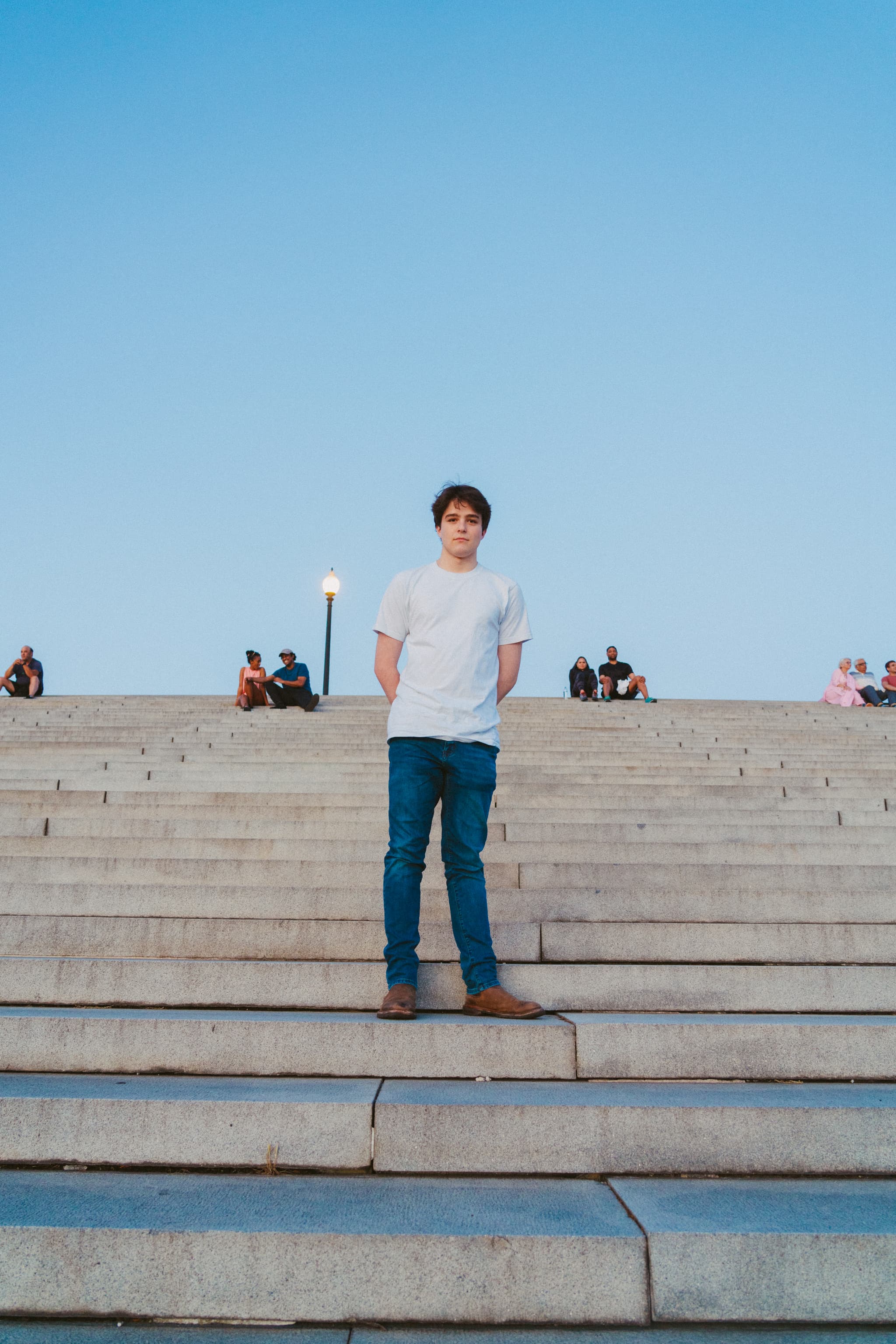 A young man stands in the center of a wide staircase with his hands slightly outstretched, wearing a white t-shirt and blue jeans. Behind him, several people are seated on the steps under a clear sky at dusk, with a street lamp glowing above his head