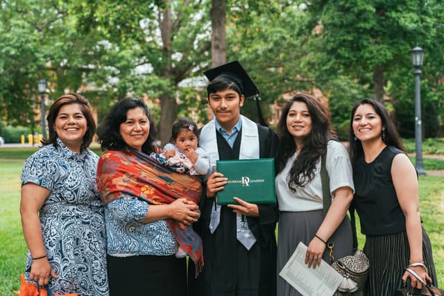 A group of five adults and a baby, presumably a family, are celebrating a graduation outdoors. The graduate is wearing a cap and gown and holding a diploma, surrounded by smiling women