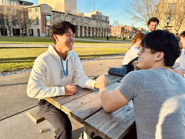 Two individuals are seated at an outdoor table, engaging in a friendly conversation, with one person smiling broadly. There's a clear sky and buildings in the background, suggesting a campus or park setting. Other people are visible in the background, indicating a social or communal area