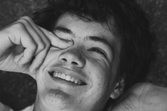 A black and white photo capturing a smiling young person lying down with one hand partially covering their face