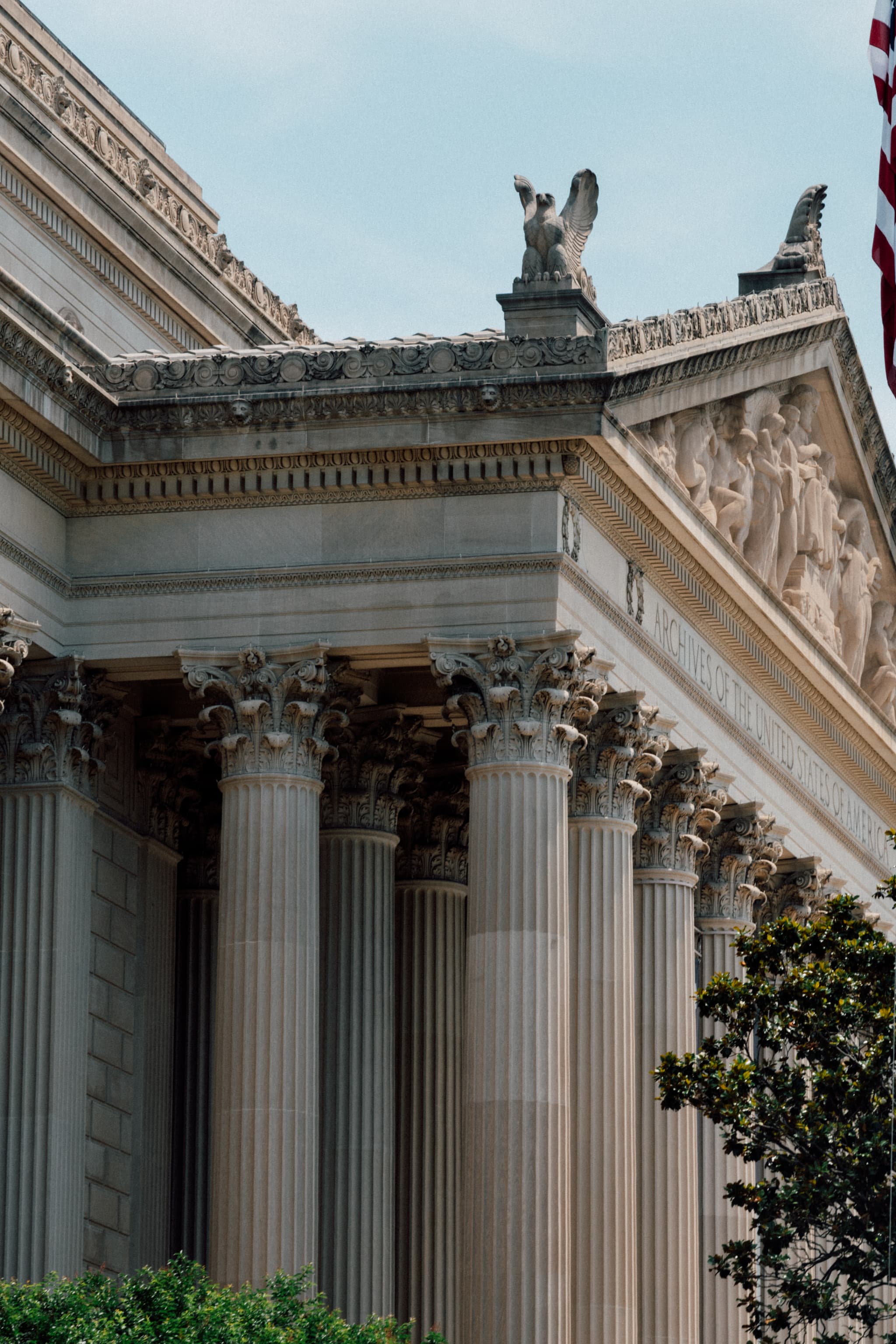 A neoclassical building facade with Corinthian columns, a sculpted pediment, and statues on the roof, partially obscured by trees, with an American flag visible to the right