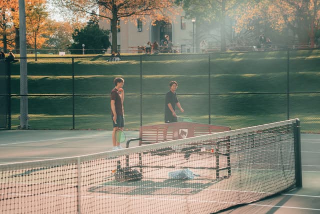 Two individuals stand on a tennis court, separated by a net, amidst a setting with trees displaying autumn foliage