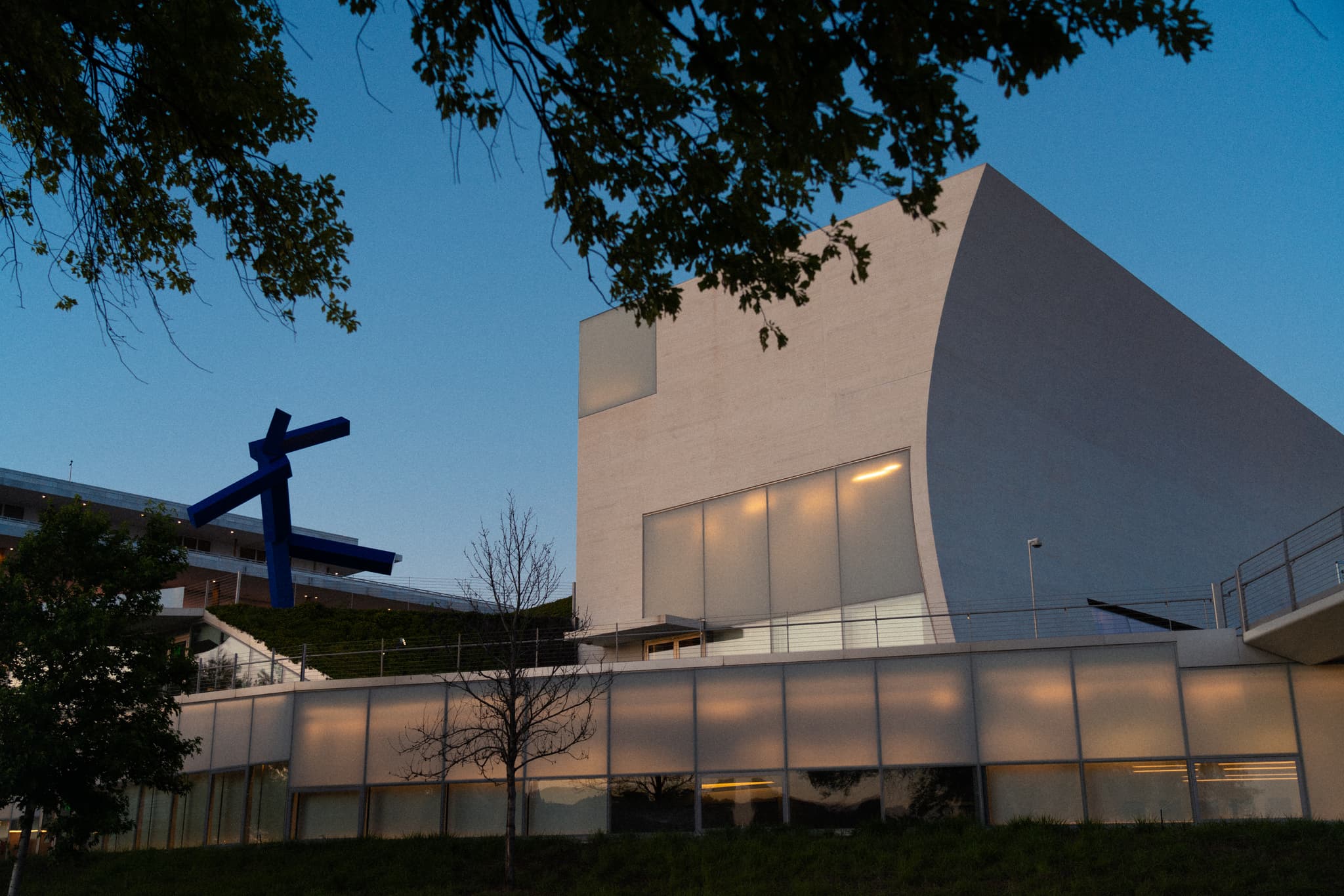 A modern building with a unique architectural design, featuring a large blue sculpture resembling an airplane on its roof, captured during twilight