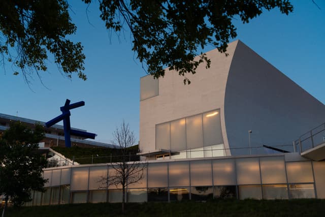 A modern building with a unique architectural design, featuring a large blue sculpture resembling an airplane on its roof, captured during twilight