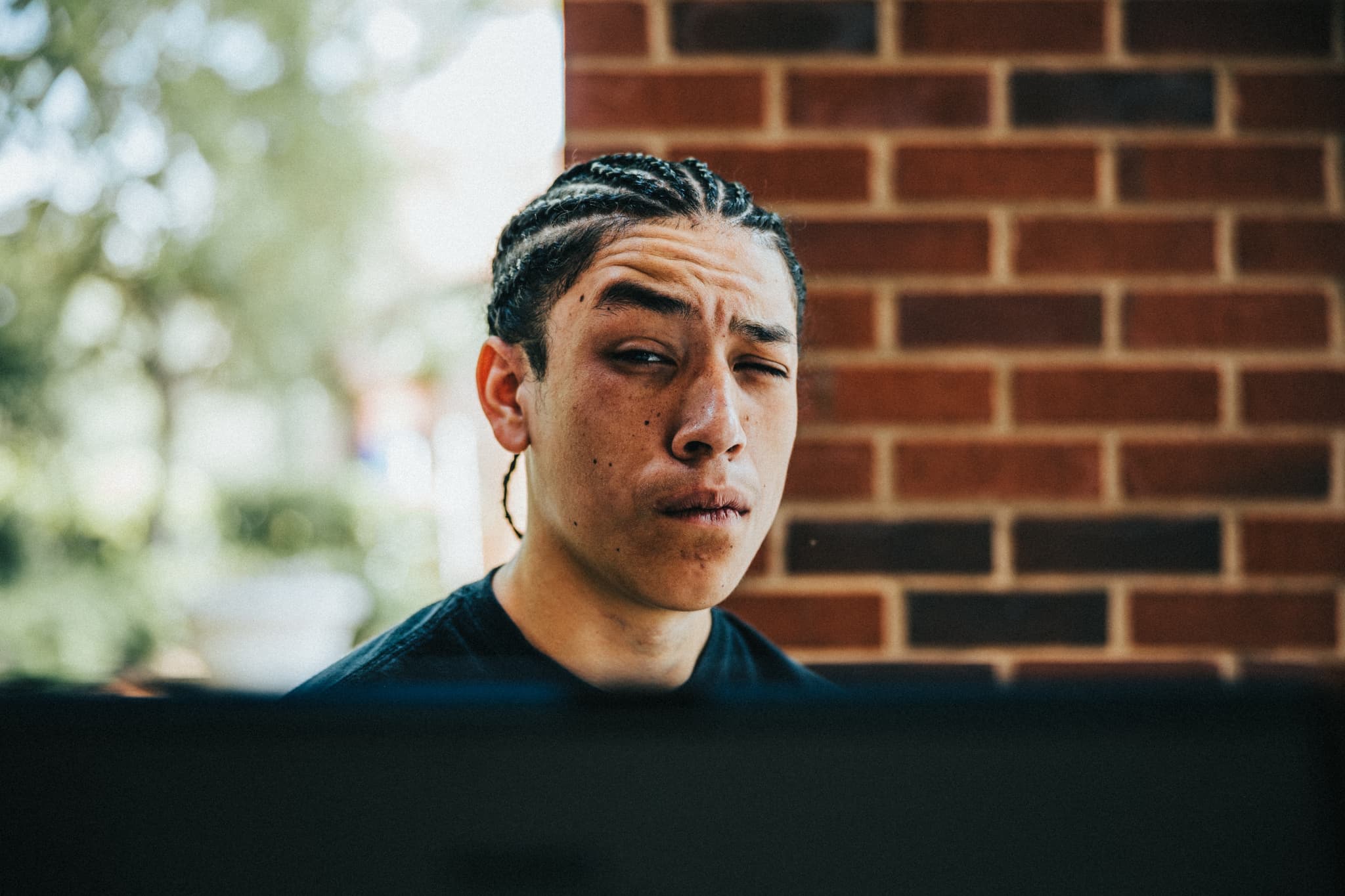 Enoch Sanchez, with braided hair appears focused and possibly looking at a computer screen, with a blurred brick wall in the background