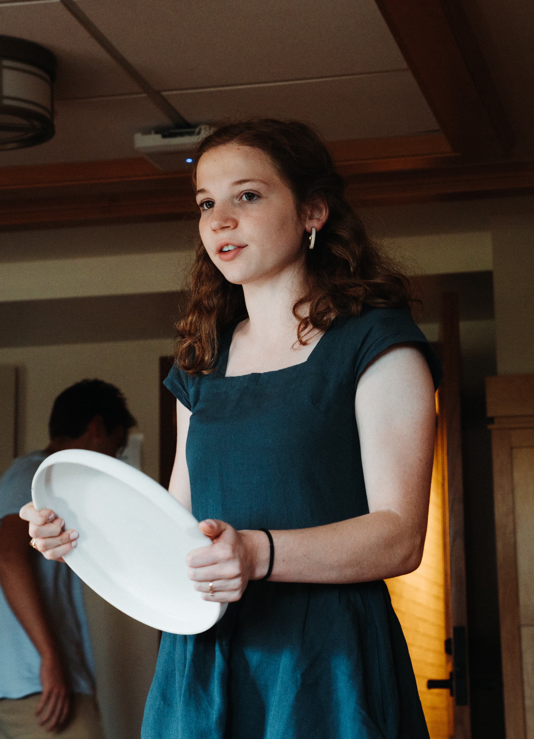 A young woman in a blue dress holds a white plate, looking to the side with a focused expression, possibly during a social event or gathering