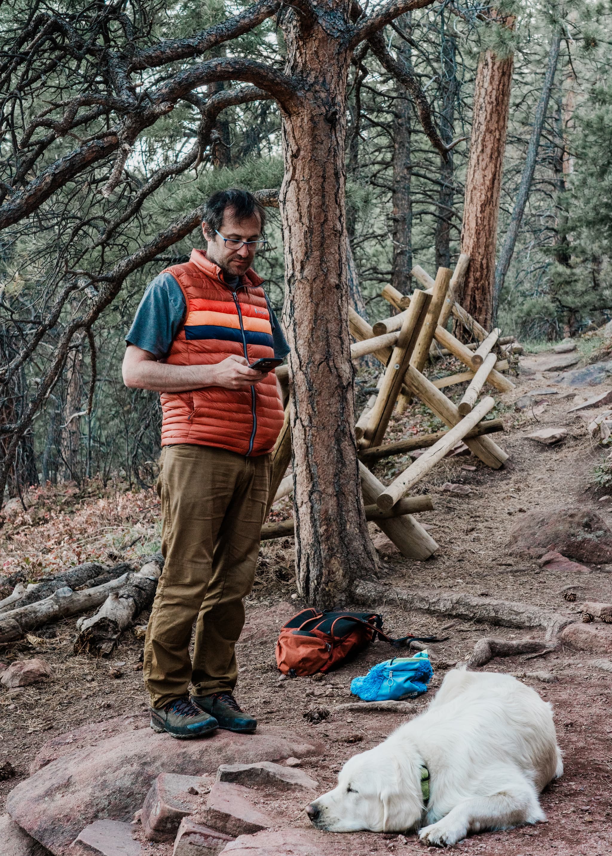 A man stands in a forested area looking at his phone, wearing a vest and glasses, with a backpack and water bottle on the ground beside him. A white dog lies resting at his feet, and there's a wooden structure in the background