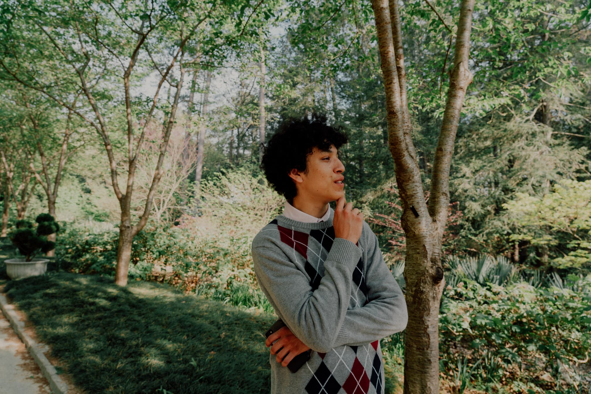 A person stands outdoors among trees, looking thoughtful with one hand on their chin