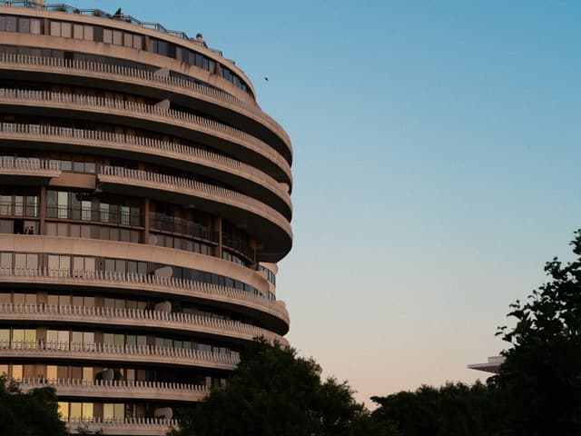 A curved, multi-story building with balconies against a twilight sky, flanked by trees