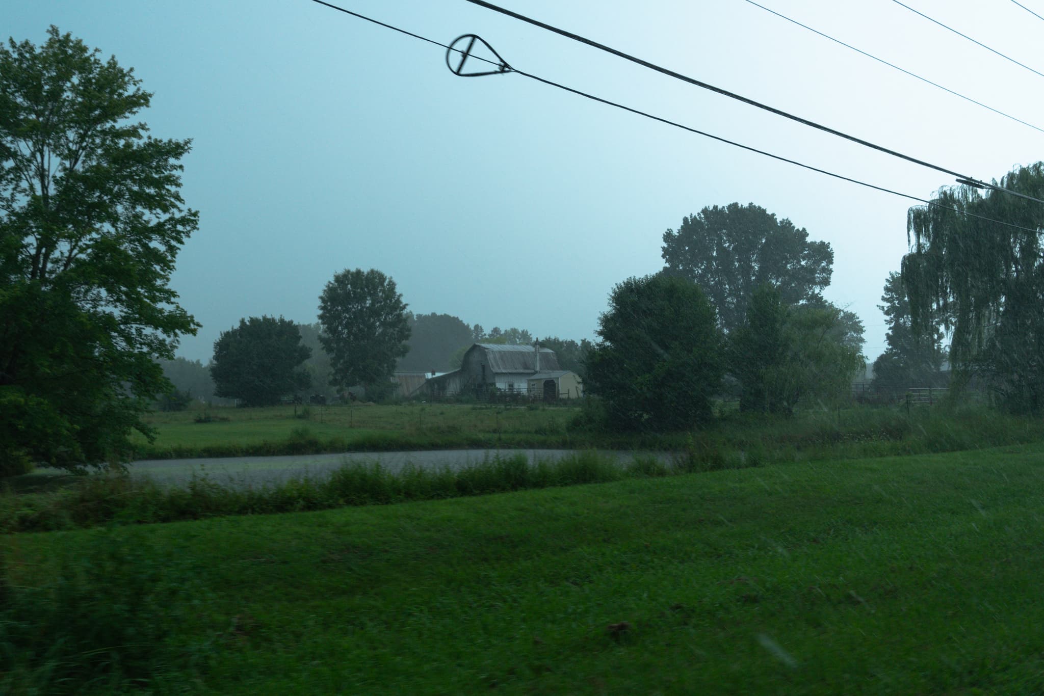 A misty rural landscape with a pond, trees, and a house in the distance, under a hazy sky with power lines crossing above