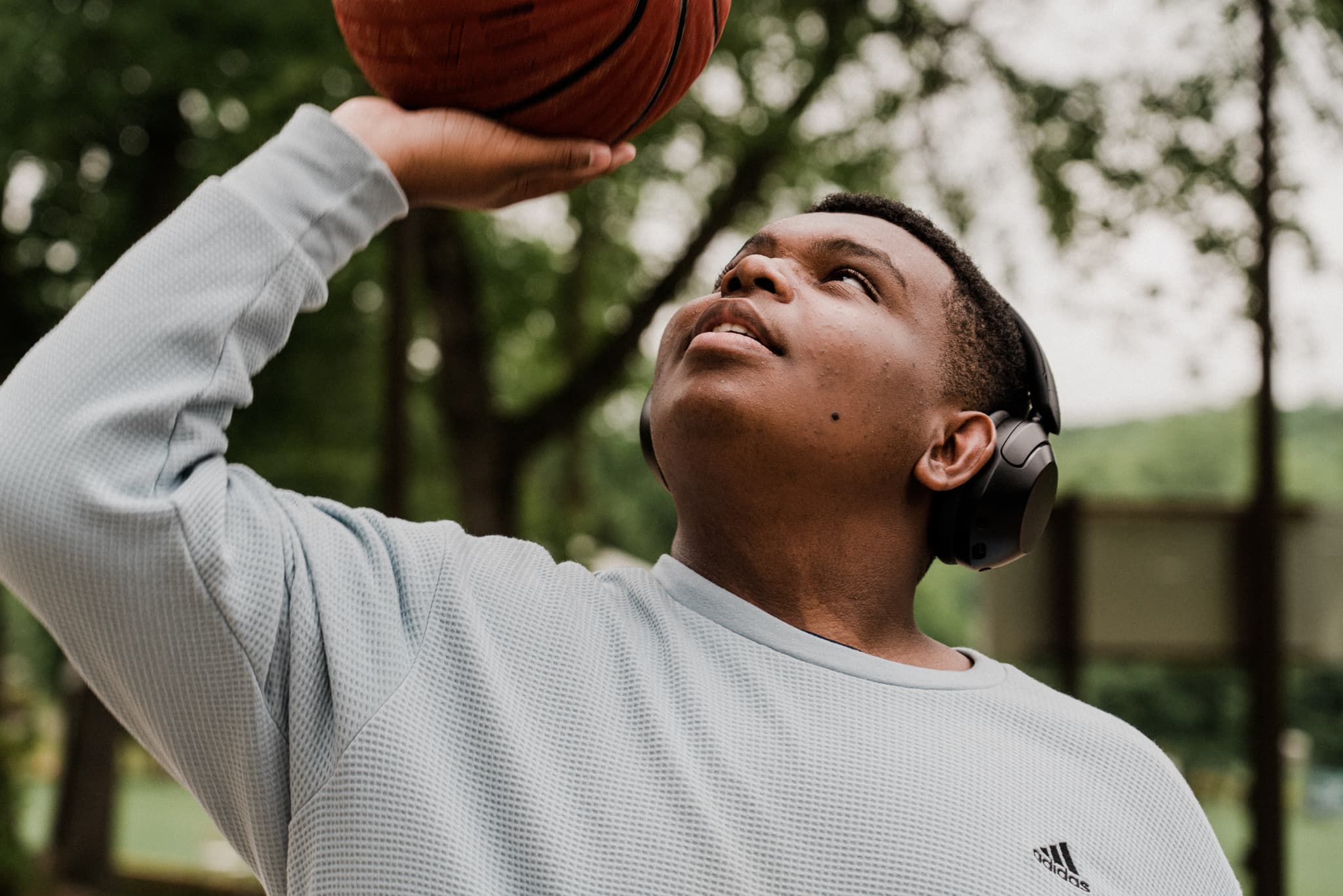 A person is focused on shooting a basketball, wearing headphones and a long-sleeve shirt, with trees in the background