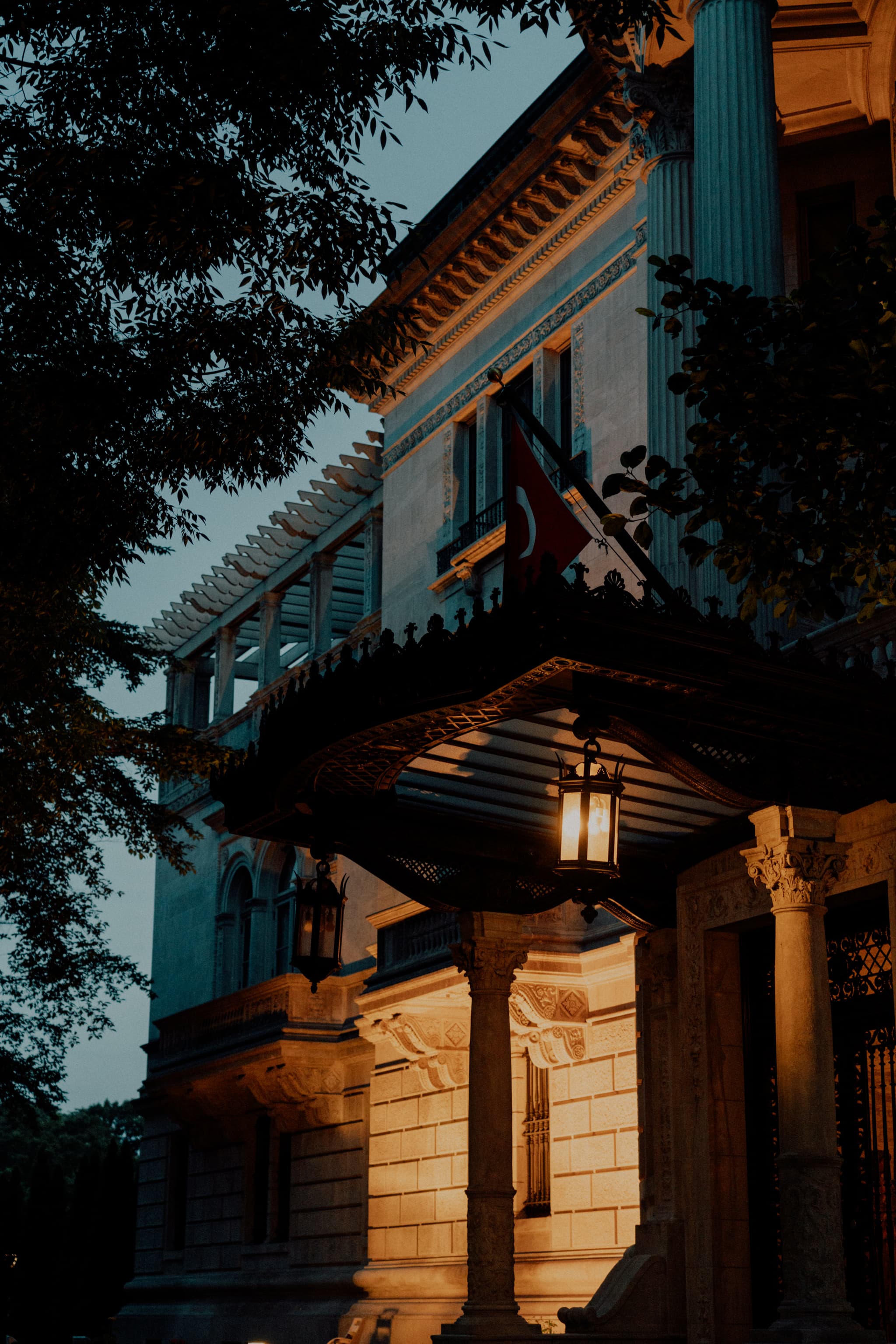 A warmly lit entrance of a classic building at dusk, featuring architectural details and a flag hanging above the doorway