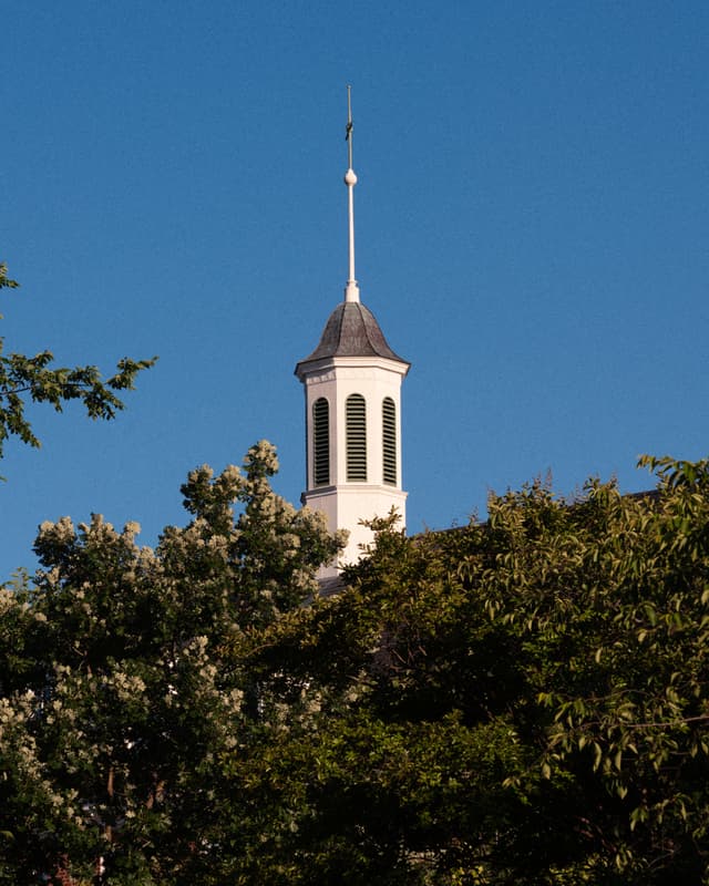 A white steeple rises above lush green trees against a clear blue sky