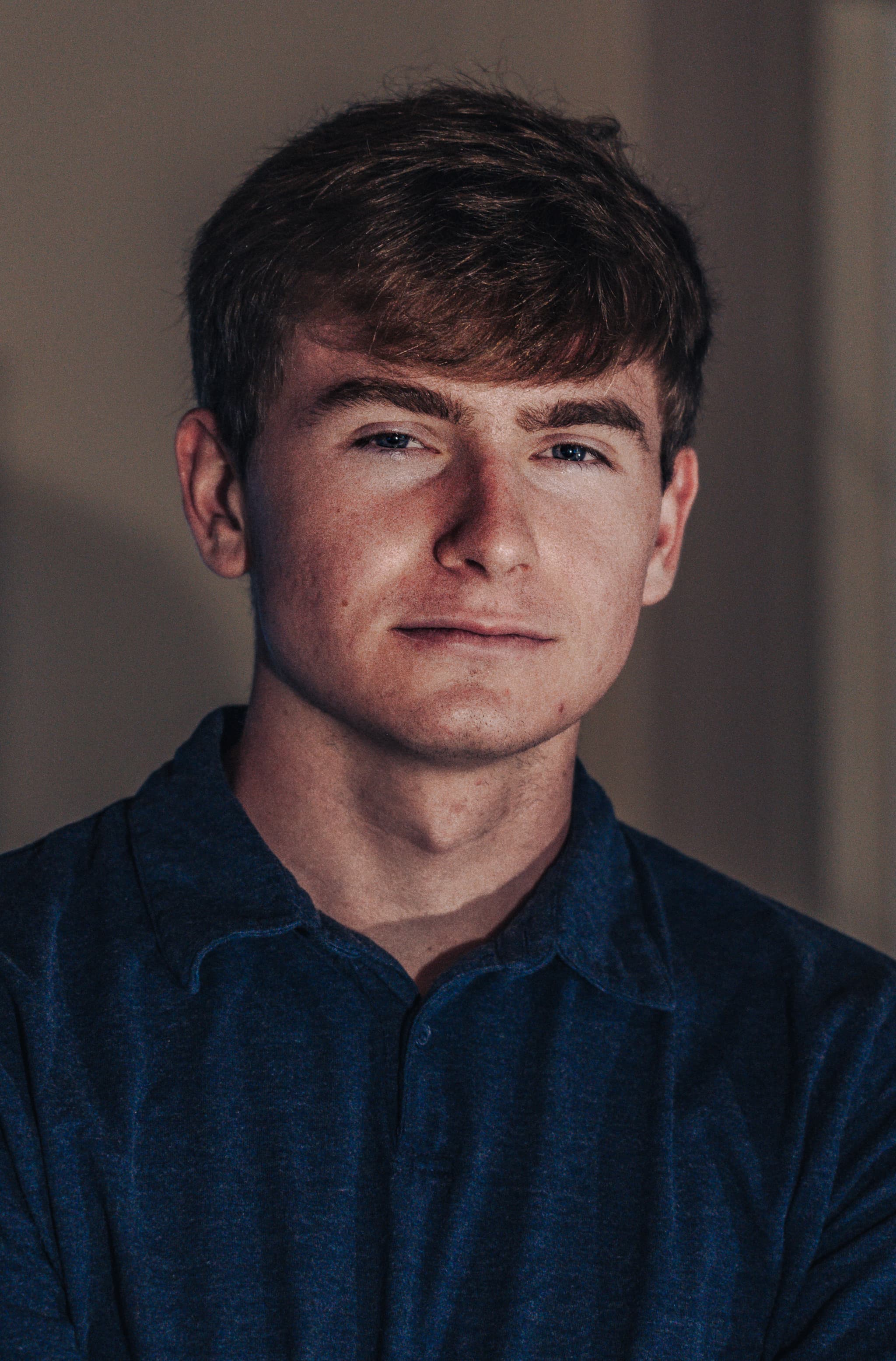 A young man with short brown hair, wearing a blue shirt, is looking directly at the camera with a slight smile. The lighting is soft and the background is blurred