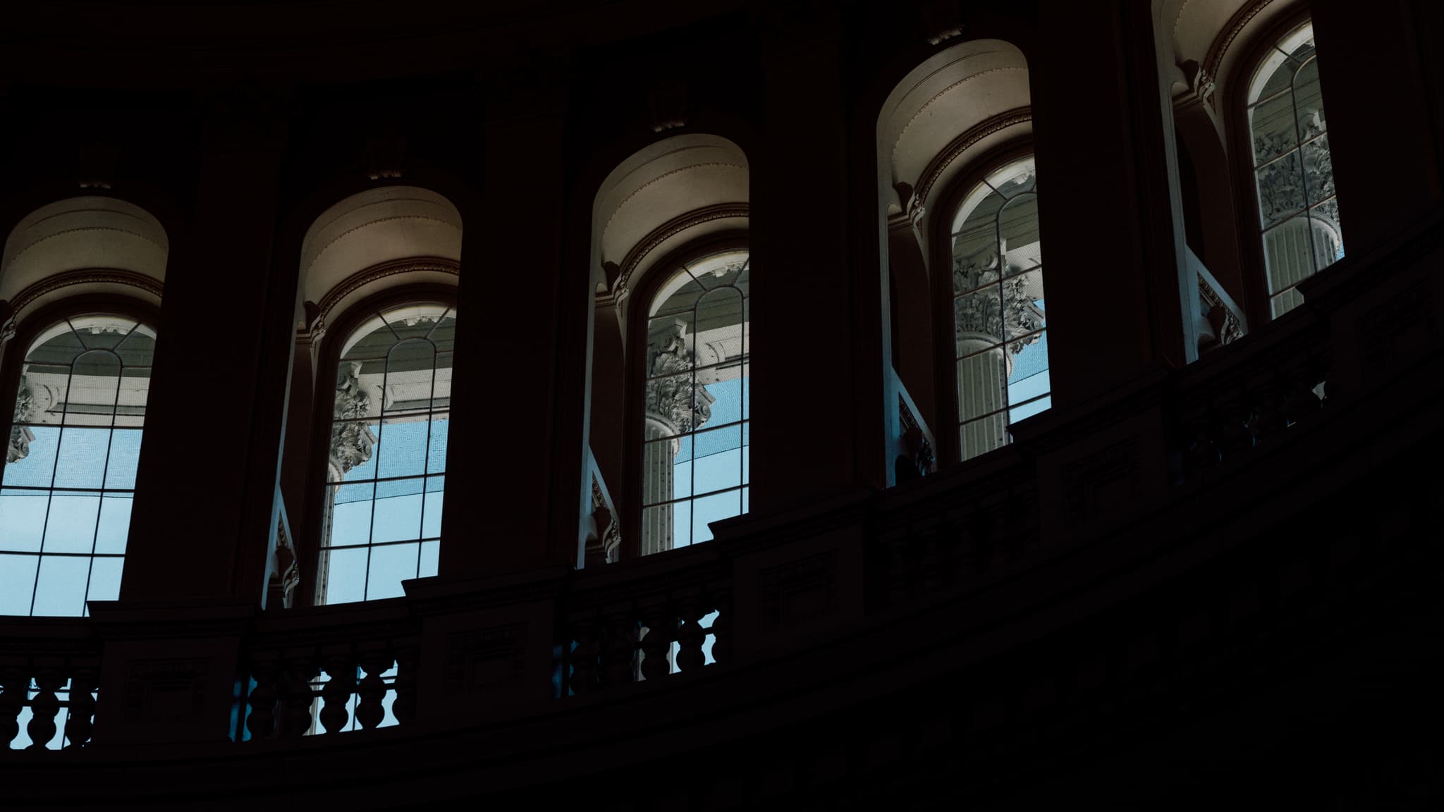 Arched windows with intricate designs cast light into a dark interior space, likely part of a grand building with a curved balcony railing visible